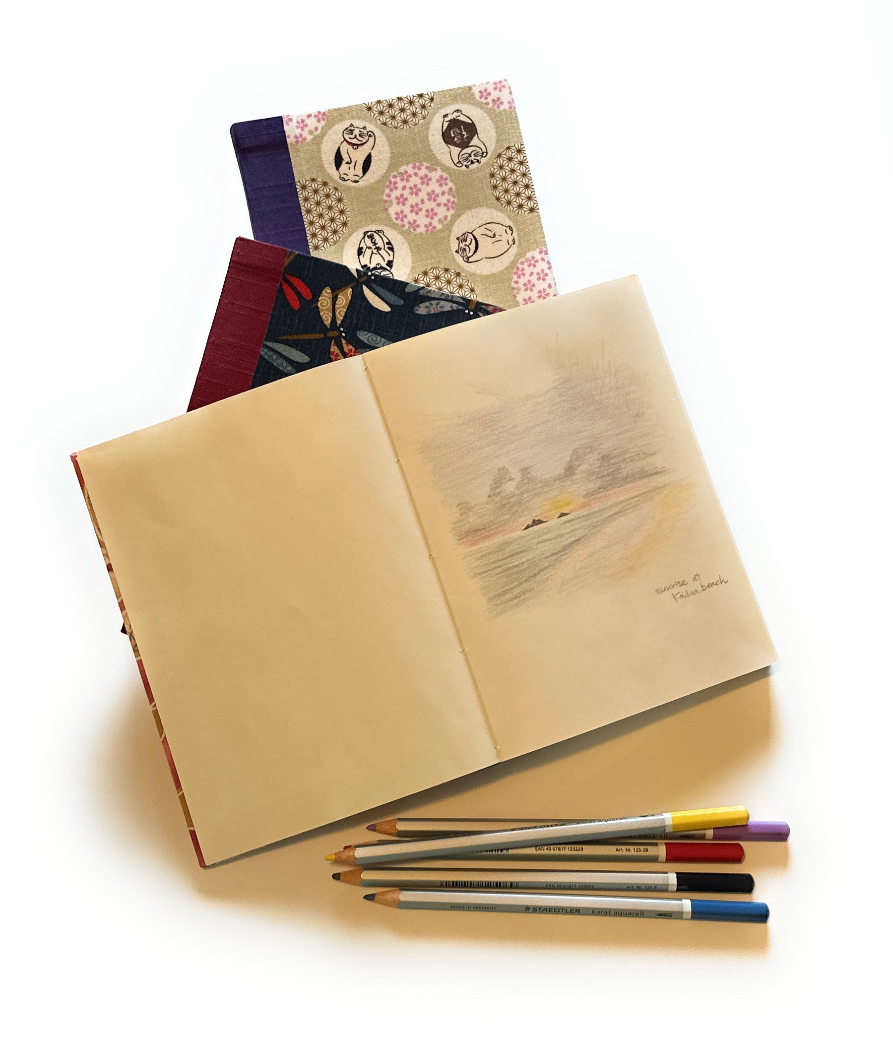An open journal displaying a soft, pastel-colored sketch titled ‘Sunrise at Kailua Beach.’ Under the journal are two other journals with beautifully patterned covers. Below the journals, a set of colored pencils is neatly arranged.