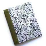 The Canopy of Leaves DIY Bookmaking Kit features a serene nature-inspired cover with delicate watercolor-style leaves in shades of green and purple on a soft white background. The journal is bound with an earthy green book cloth spine, creating an elegant and organic aesthetic. This DIY kit includes all the materials needed to craft a handmade booklet, making it a perfect creative project for journal lovers, writers, and stationery enthusiasts who appreciate artisan craftsmanship and natural beauty.