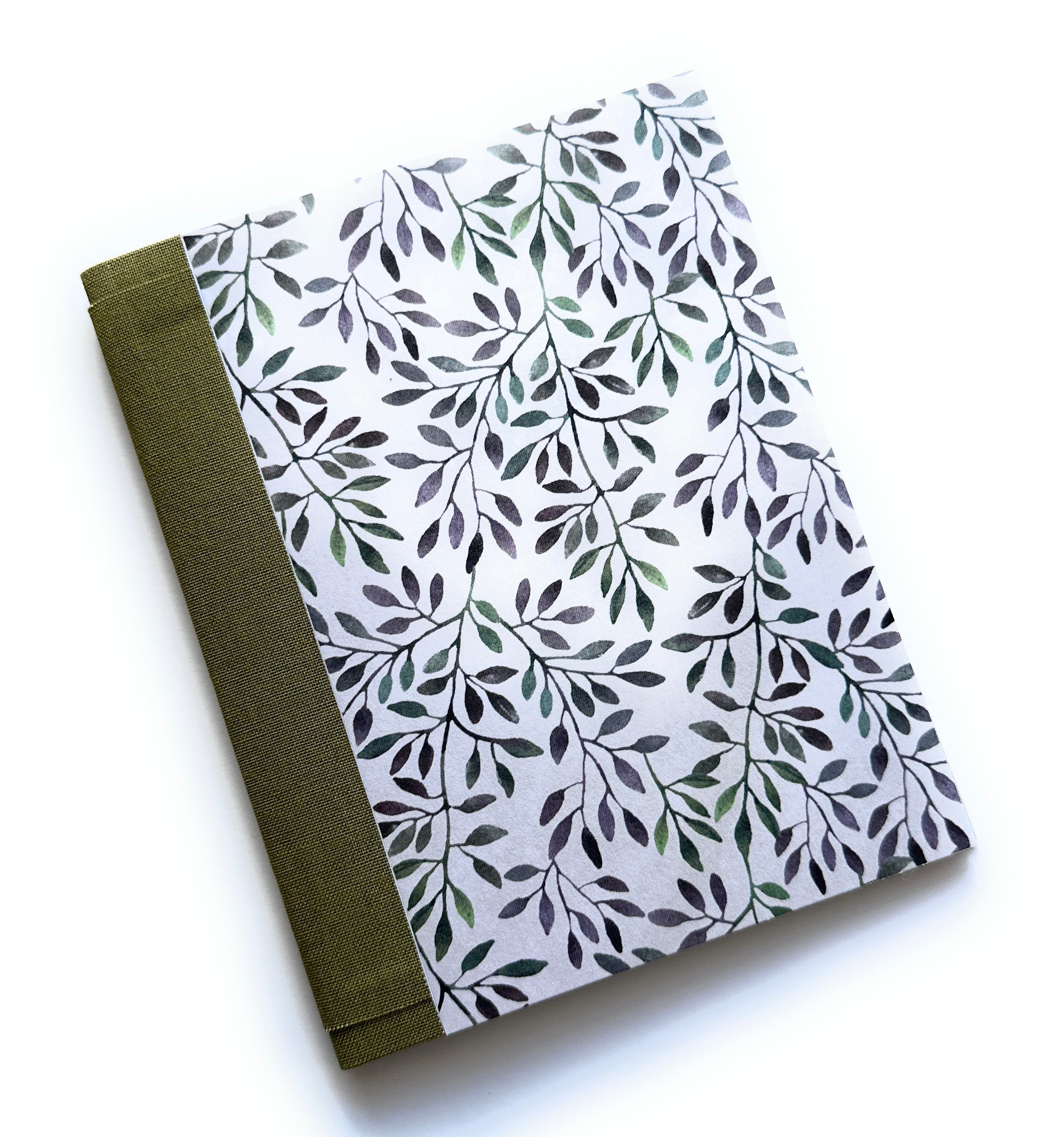 The Canopy of Leaves DIY Bookmaking Kit features a serene nature-inspired cover with delicate watercolor-style leaves in shades of green and purple on a soft white background. The journal is bound with an earthy green book cloth spine, creating an elegant and organic aesthetic. This DIY kit includes all the materials needed to craft a handmade booklet, making it a perfect creative project for journal lovers, writers, and stationery enthusiasts who appreciate artisan craftsmanship and natural beauty.