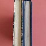 A close-up view of two handmade journals side by side, showcasing their detailed spines. One features a tropical botanical fabric cover, while the other has a sleek navy blue cloth cover, highlighting the craftsmanship and unique binding style.