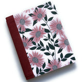 The Daisy Dreams DIY Bookmaking Kit features a beautifully handcrafted cover adorned with soft pink daisies and deep green leaves on a light background. Complemented by a rich burgundy book cloth spine, this elegant DIY kit provides all the materials needed to create a personalized handmade book. Ideal for journal enthusiasts, writers, and craft lovers, this bookbinding kit offers a unique and creative way to make a custom floral-themed notebook.