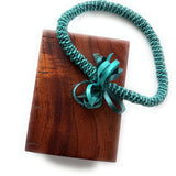 A handcrafted Koa wood Coptic journal featuring a smooth, polished finish. A teal ribbon lei with intricate woven details and a decorative bow is draped over the journal.