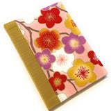 This handmade journal features a vibrant cherry blossom fabric cover with red, gold, white, and purple floral patterns set against a soft pink background. The spine is wrapped in a textured golden bookcloth, adding an elegant contrast. Crafted with high-quality materials, this Japanese-inspired notebook is ideal for writing, journaling, or sketching. A perfect gift for stationery lovers, writers, and artists, this unique piece blends traditional craftsmanship with modern functionality.