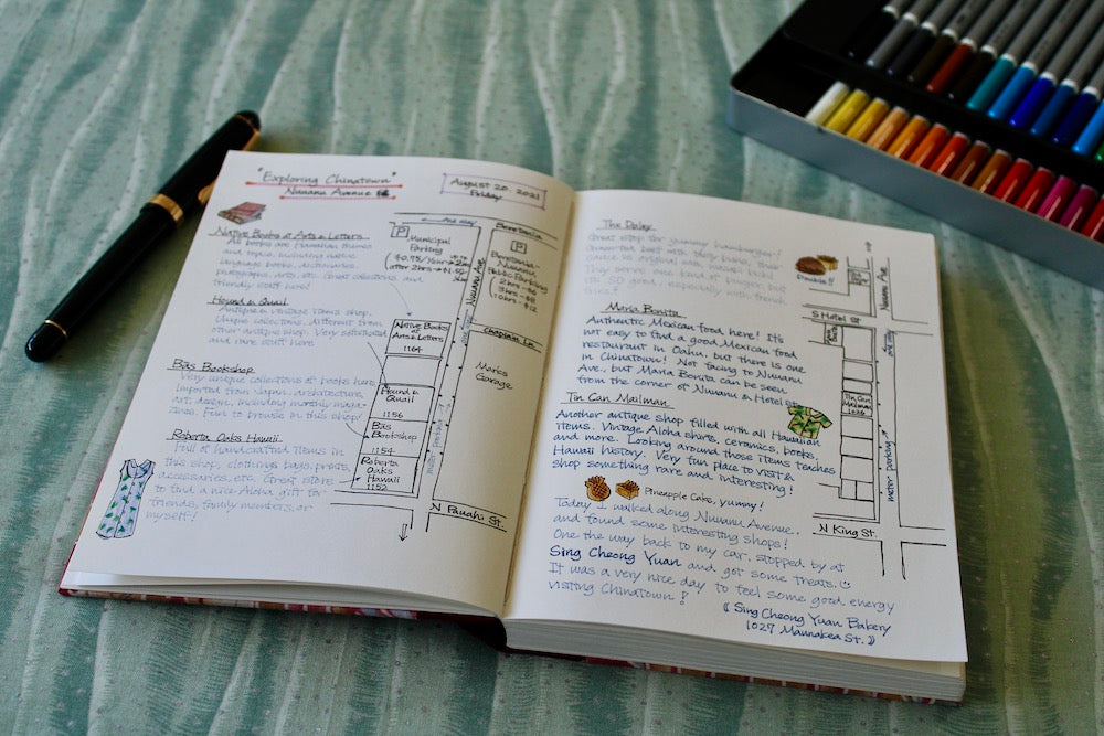 An open journal with handwritten notes and sketches lies on a light green fabric surface. The journal features a detailed exploration of Honolulu’s Chinatown, including maps, shop descriptions, and personal reflections. A black fountain pen is placed beside the journal, and a box of colorful pens or markers is partially visible in the background. The journal pages are neatly organized with headings, bullet points, and small illustrations, creating an engaging and visually appealing layout.