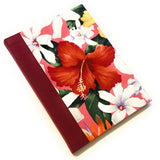 This image shows a handmade book with a vibrant, tropical fabric cover. The cover features large, colorful flowers, including a prominent red hibiscus, Tahitian gardenias, orchids, and other tropical blooms set against a pink background. The spine is covered with a deep red fabric that complements the bright floral design. The book is expertly crafted, exhibiting a clean and polished finish.