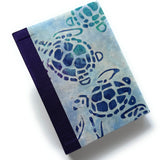 A handmade journal featuring a batik-style cover with blue and green sea turtles (“honu”) swimming across a soft watercolor background, paired with a deep indigo spine.