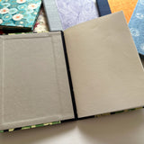 An open handmade keepsake card displaying high-quality textured pages and a fabric-covered hardcover. Perfect for personalized messages, well-wishes, or special occasions, this booklet provides ample space for writing while ensuring a beautifully crafted presentation. Surrounded by other unique handcrafted designs.