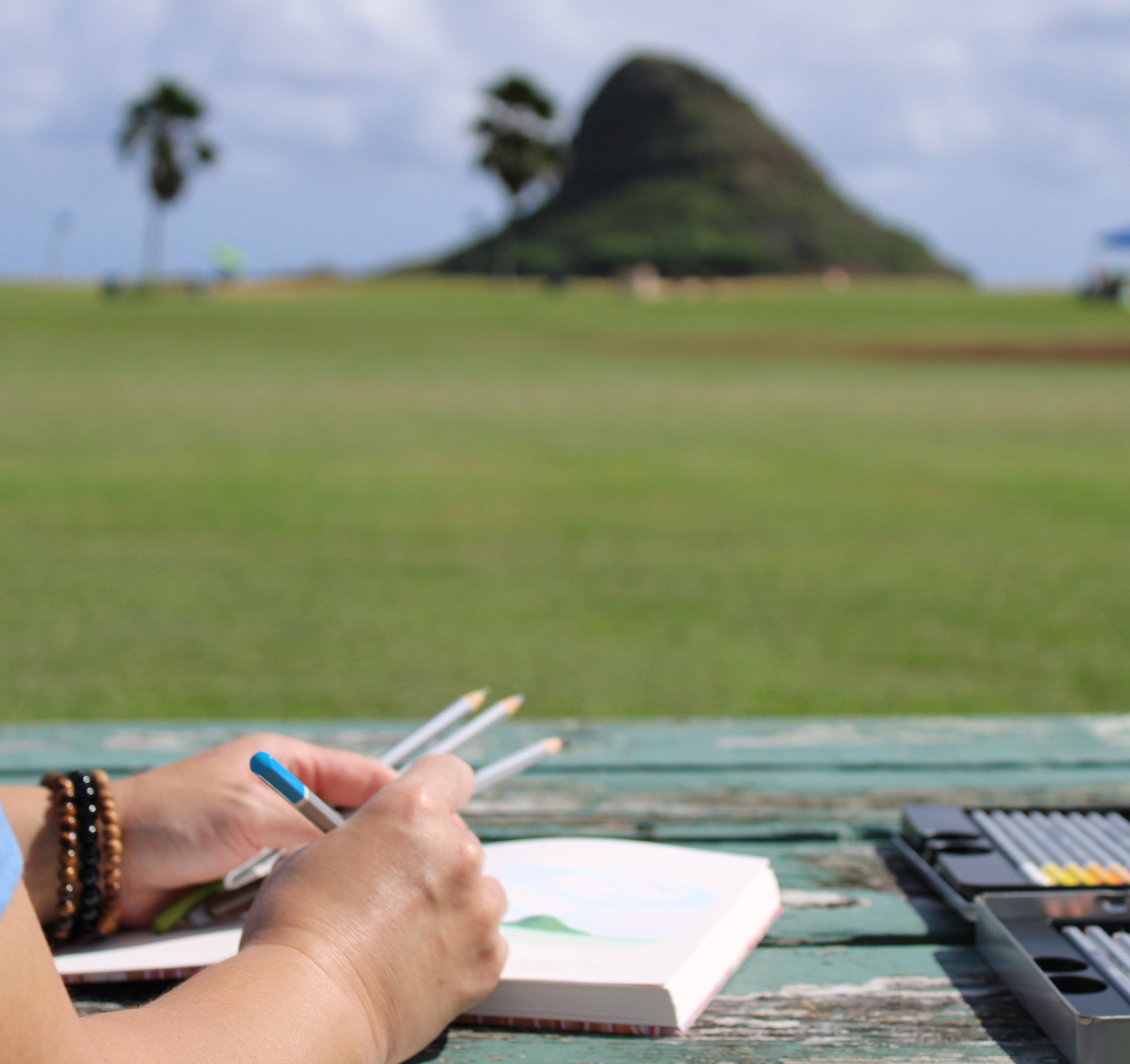 A sketchbook journaler captures the scenic beauty of Mokoliʻi (Chinaman’s Hat) in Hawaiʻi, seated at a rustic picnic table with colored pencils and an open notebook. The image highlights the art of outdoor sketching, travel journaling, and creative inspiration, set against a backdrop of lush green fields, palm trees, and a clear blue sky. Ideal for artists, travel enthusiasts, and nature lovers seeking to document experiences through hand-drawn illustrations and mindful creativity.