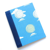 The World Awaits DIY Bookmaking Kit features a vibrant blue cover adorned with illustrated globes and fluffy white clouds, evoking a sense of adventure and exploration. Bound with a deep navy blue book cloth spine, this handmade journal kit is perfect for travelers, writers, and creatives looking to document their journeys or dreams. The kit includes all the materials needed to craft a personalized booklet, making it an ideal project for bookbinding enthusiasts and those who love unique, travel-themed stati