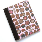 The Truffle Tales DIY Bookmaking Kit features a delightful chocolate-themed cover design with truffles, bonbons, and sweet treats on a pastel pink background. This handcrafted pocket journal includes a dark brown book cloth spine and is part of a creative craft kit perfect for book lovers, writers, and DIY enthusiasts looking to create their own personalized booklet. Ideal as a thoughtful gift or a fun project for chocolate and stationery fans alike.