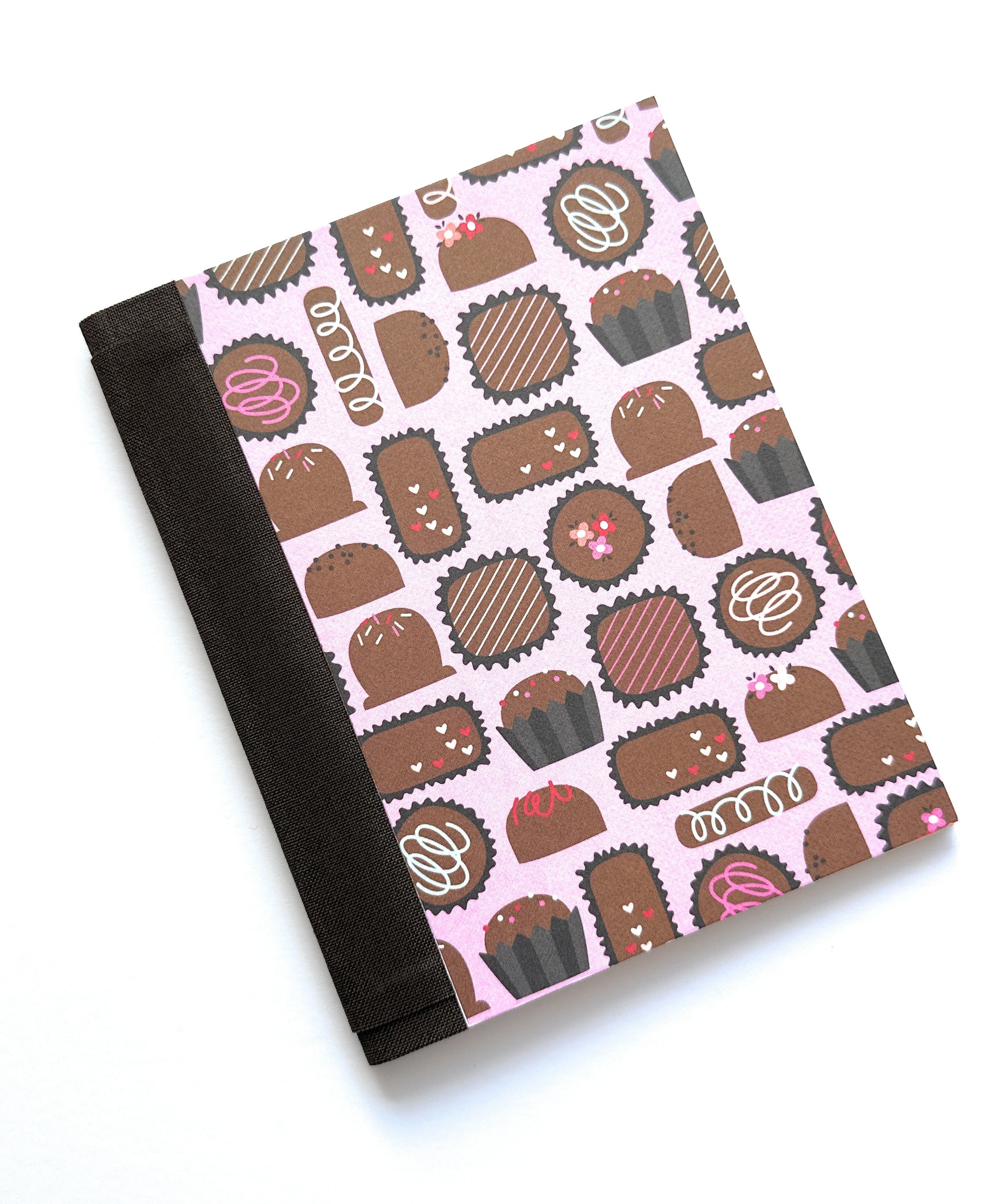 The Truffle Tales DIY Bookmaking Kit features a delightful chocolate-themed cover design with truffles, bonbons, and sweet treats on a pastel pink background. This handcrafted pocket journal includes a dark brown book cloth spine and is part of a creative craft kit perfect for book lovers, writers, and DIY enthusiasts looking to create their own personalized booklet. Ideal as a thoughtful gift or a fun project for chocolate and stationery fans alike.