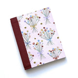 The Wildflower Whimsy DIY Bookmaking Kit features a charming pastel pink cover decorated with delicate bouquets of wildflowers tied with ribbons, accompanied by tiny buzzing bees. The book’s deep burgundy book cloth spine adds an elegant contrast, making it a perfect journal for nature lovers, writers, and creatives. This DIY bookbinding kit includes all the materials needed to craft a handmade booklet, offering a delightful project for those who appreciate floral-inspired stationery and personalized notebo