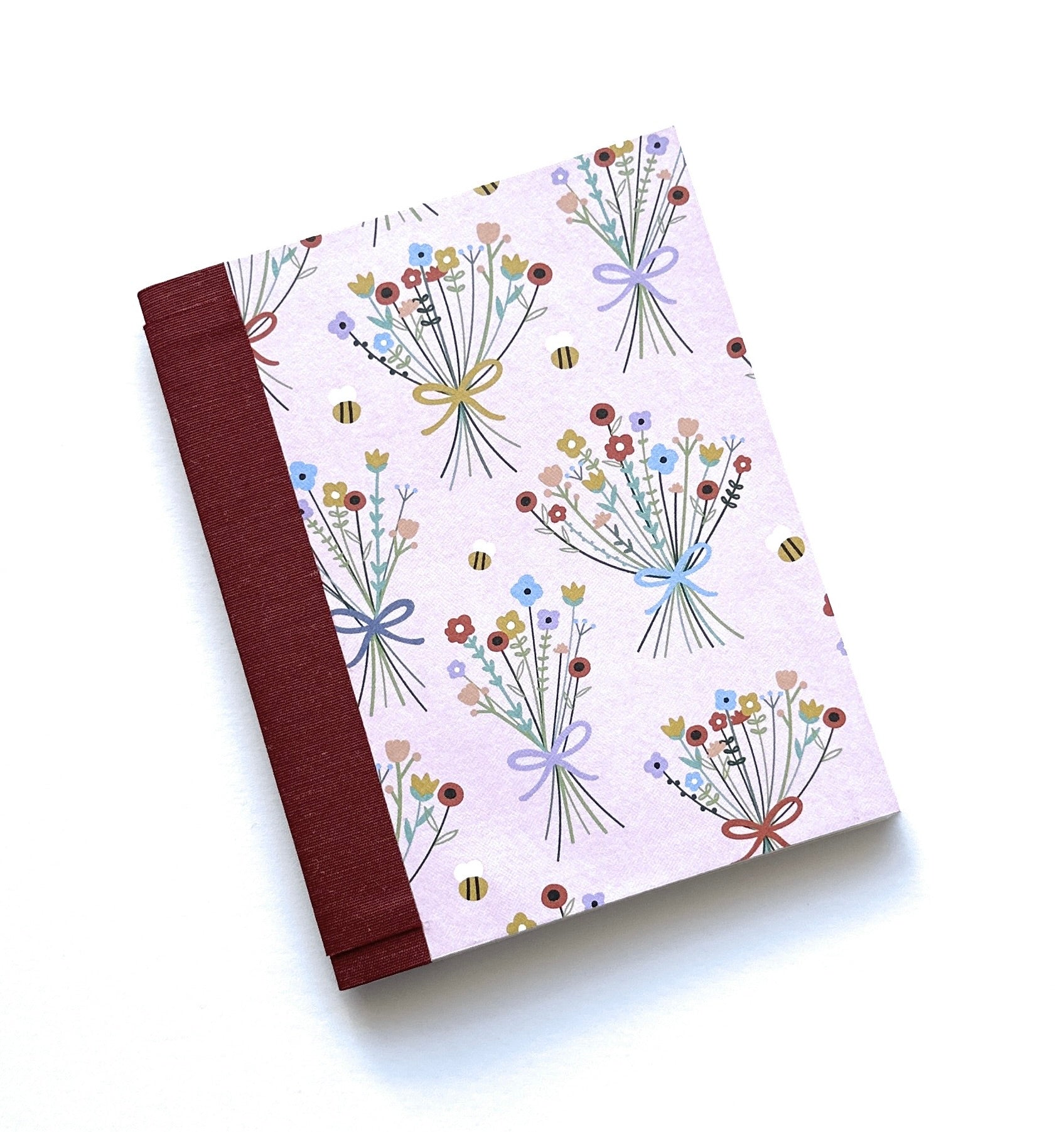 The Wildflower Whimsy DIY Bookmaking Kit features a charming pastel pink cover decorated with delicate bouquets of wildflowers tied with ribbons, accompanied by tiny buzzing bees. The book’s deep burgundy book cloth spine adds an elegant contrast, making it a perfect journal for nature lovers, writers, and creatives. This DIY bookbinding kit includes all the materials needed to craft a handmade booklet, offering a delightful project for those who appreciate floral-inspired stationery and personalized notebo