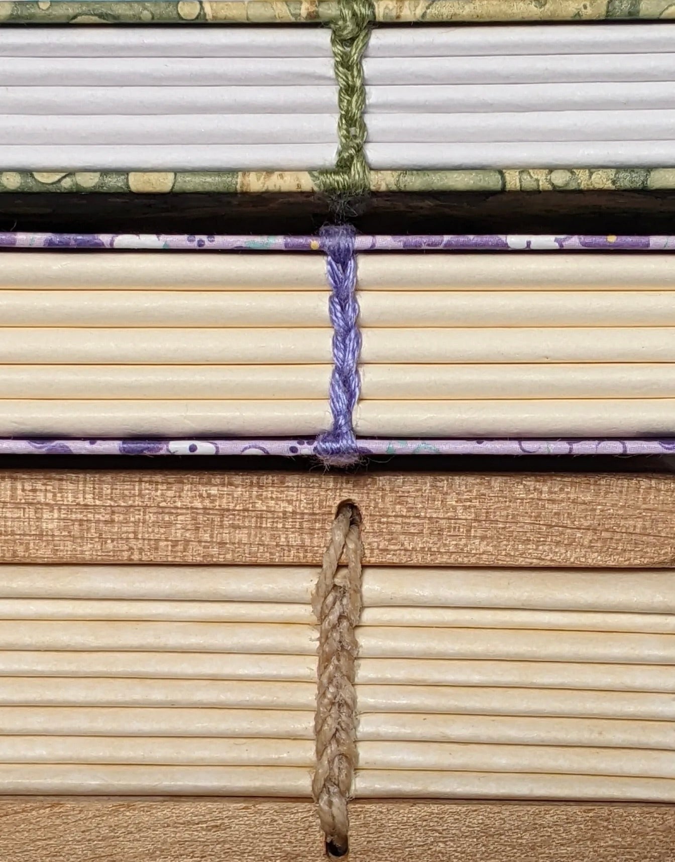 A close-up view of the spines of three handmade books stacked on top of each other, showcasing different binding techniques. The top book has a green marbled cover with green thread binding, the middle book has a purple and white patterned cover with purple thread binding, and the bottom book has a golden cover with beige thread binding. The intricate stitching on each book’s spine highlights the craftsmanship and variety in bookbinding methods.