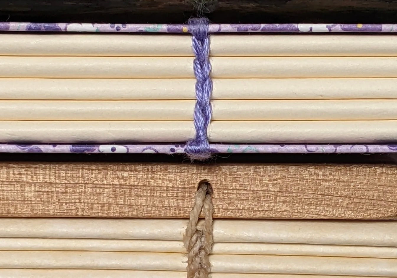 A close-up view of the spines of three handmade books stacked on top of each other, showcasing different binding techniques. The top book has a green marbled cover with green thread binding, the middle book has a purple and white patterned cover with purple thread binding, and the bottom book has a golden cover with beige thread binding. The intricate stitching on each book’s spine highlights the craftsmanship and variety in bookbinding methods.