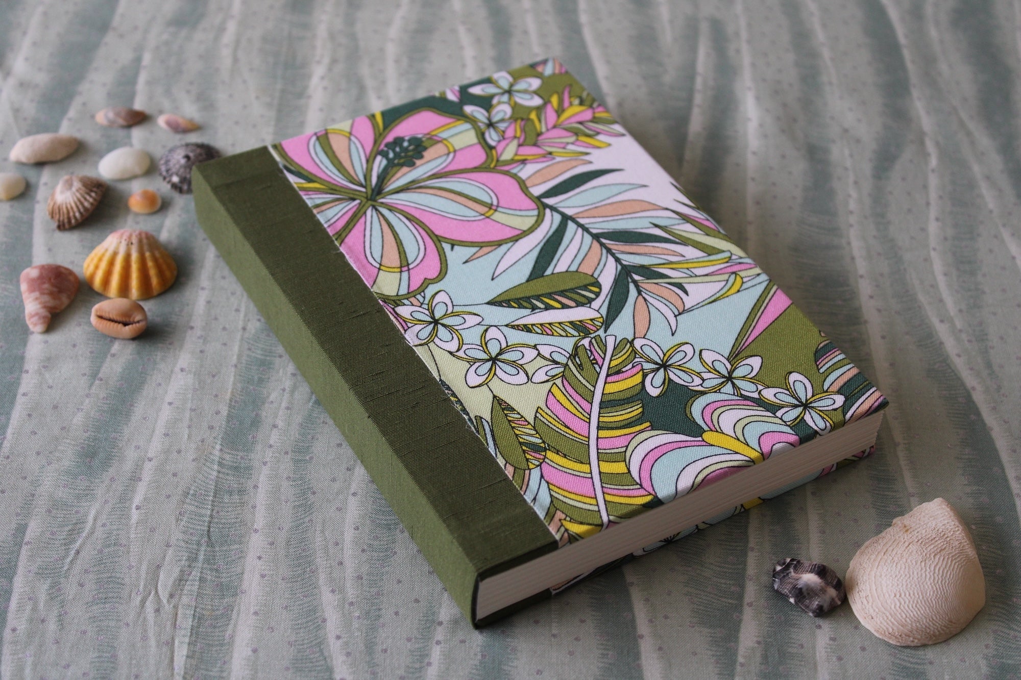Handmade journal from Kuro’s Workshop featuring a Hawaiian-inspired floral fabric cover with green bookcloth spine, displayed with seashells on a textured fabric background.