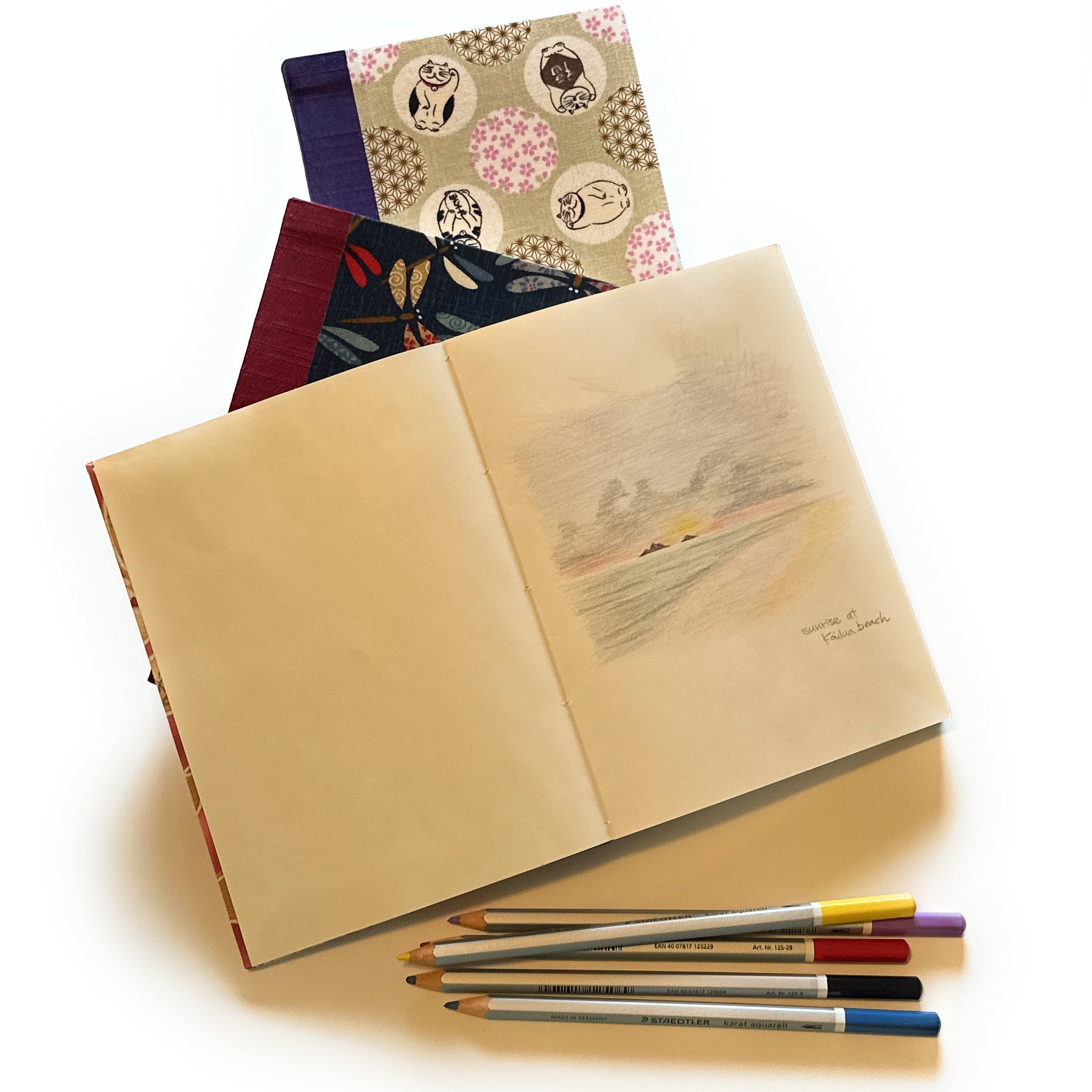 An open journal displaying a soft, pastel-colored sketch titled ‘Sunrise at Kailua Beach.’ Under the journal are two other journals with beautifully patterned covers. Below the journals, a set of colored pencils is neatly arranged.