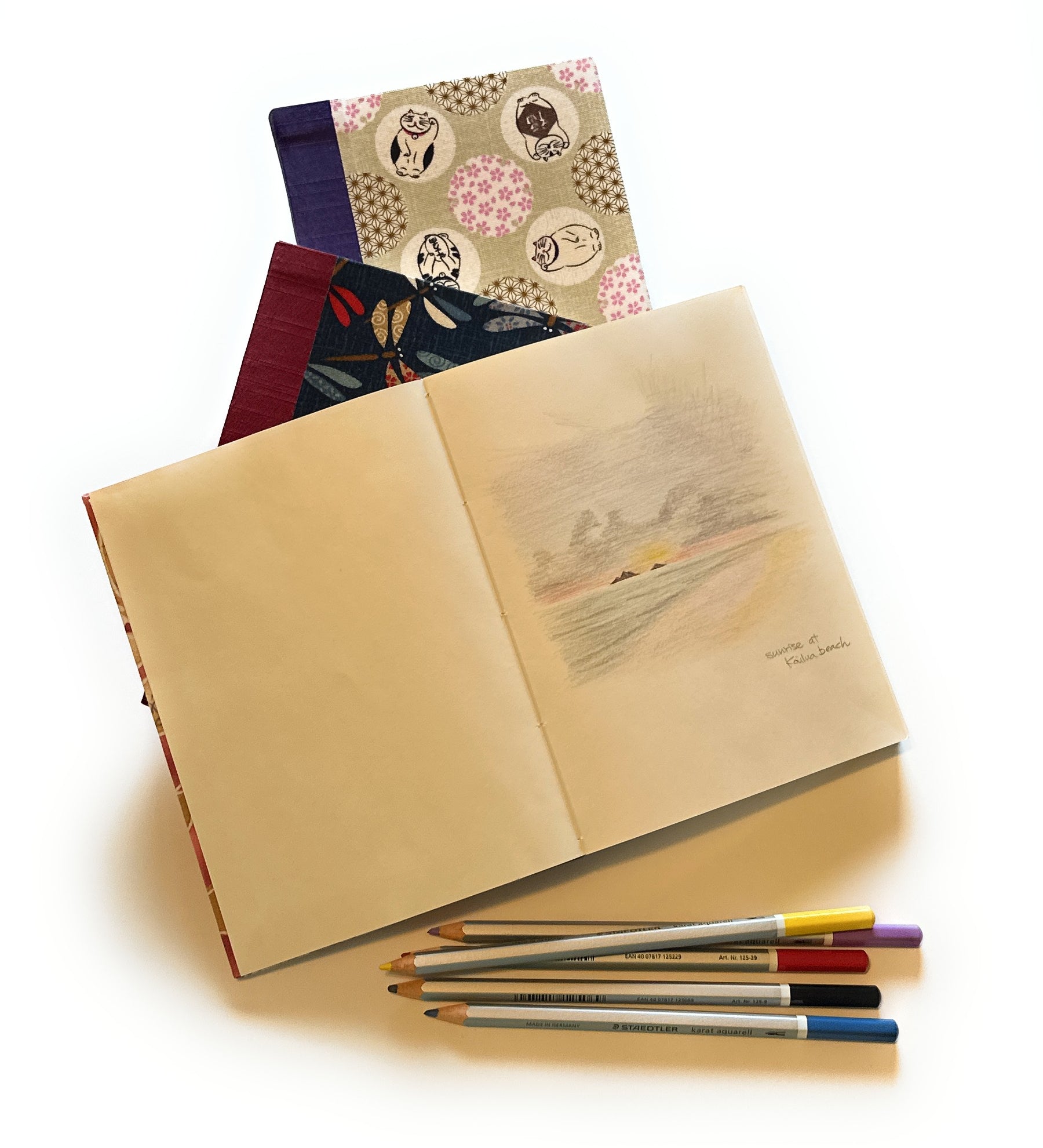 An open journal displaying a soft, pastel-colored sketch titled ‘Sunrise at Kailua Beach.’ Under the journal are two other journals with beautifully patterned covers. Below the journals, a set of colored pencils is neatly arranged.