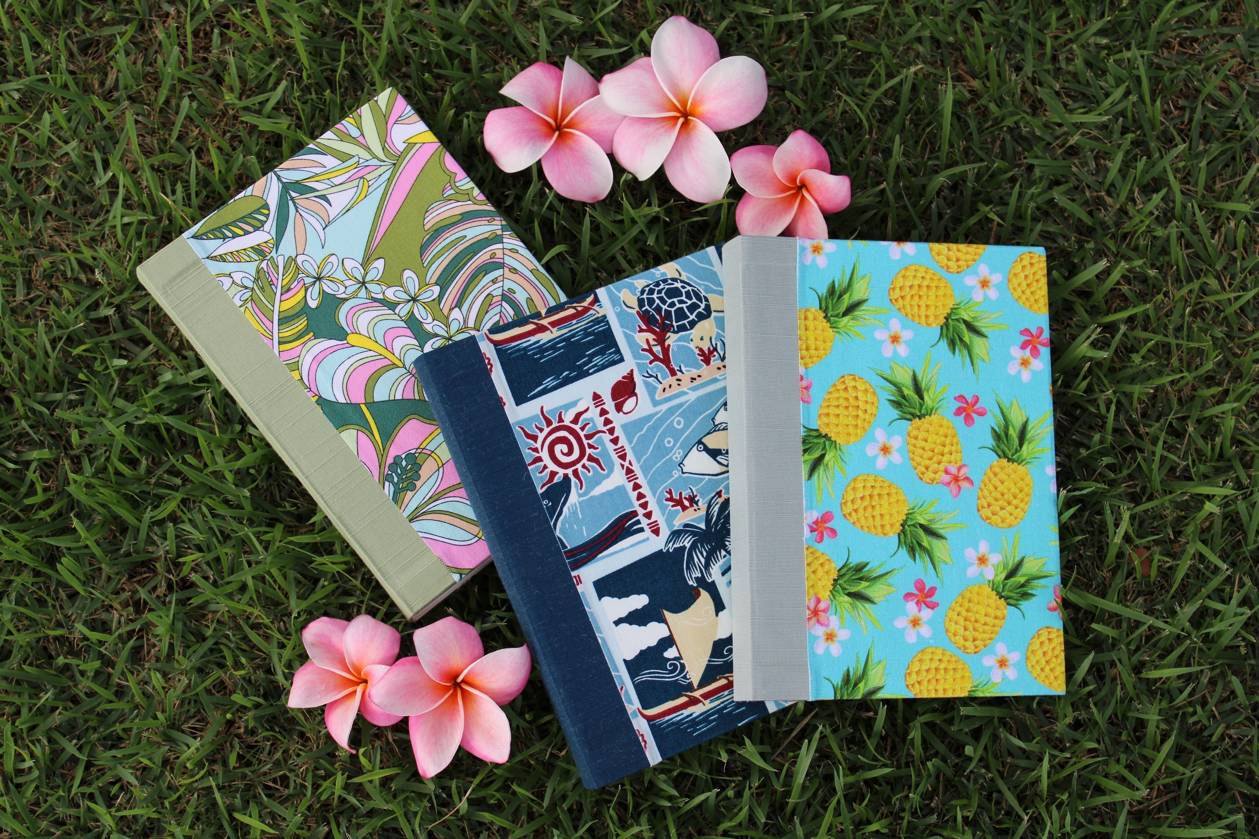 Three Aloha Journals on green grass surrounded by pink plumerias.