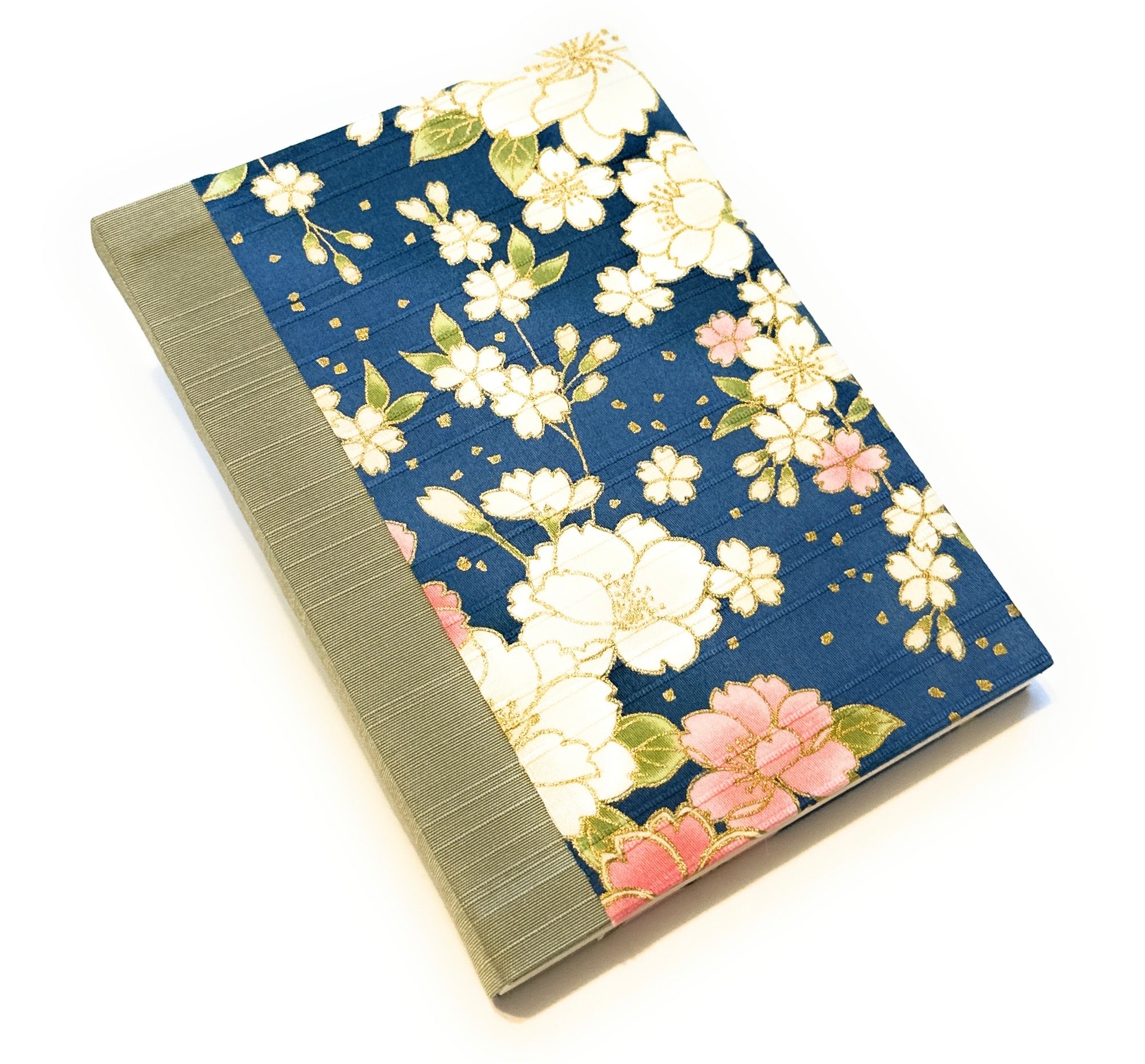 This image shows a handmade book with a dark blue fabric cover featuring a floral design. The cover is adorned with large white flowers, smaller pink flowers, and green leaves, all accented with gold details. The spine is covered with a light olive-green fabric, providing a subtle contrast to the vibrant floral pattern. The book appears to be meticulously crafted, with a clean and professional finish.