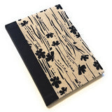 This image shows a handmade book with a beige fabric cover featuring a minimalist black floral pattern. The design includes various black flowers and vertical lines that create a natural, organic look. The spine is covered with solid black fabric, providing a striking contrast to the patterned cover. The book is skillfully crafted, showcasing a clean and professional finish.