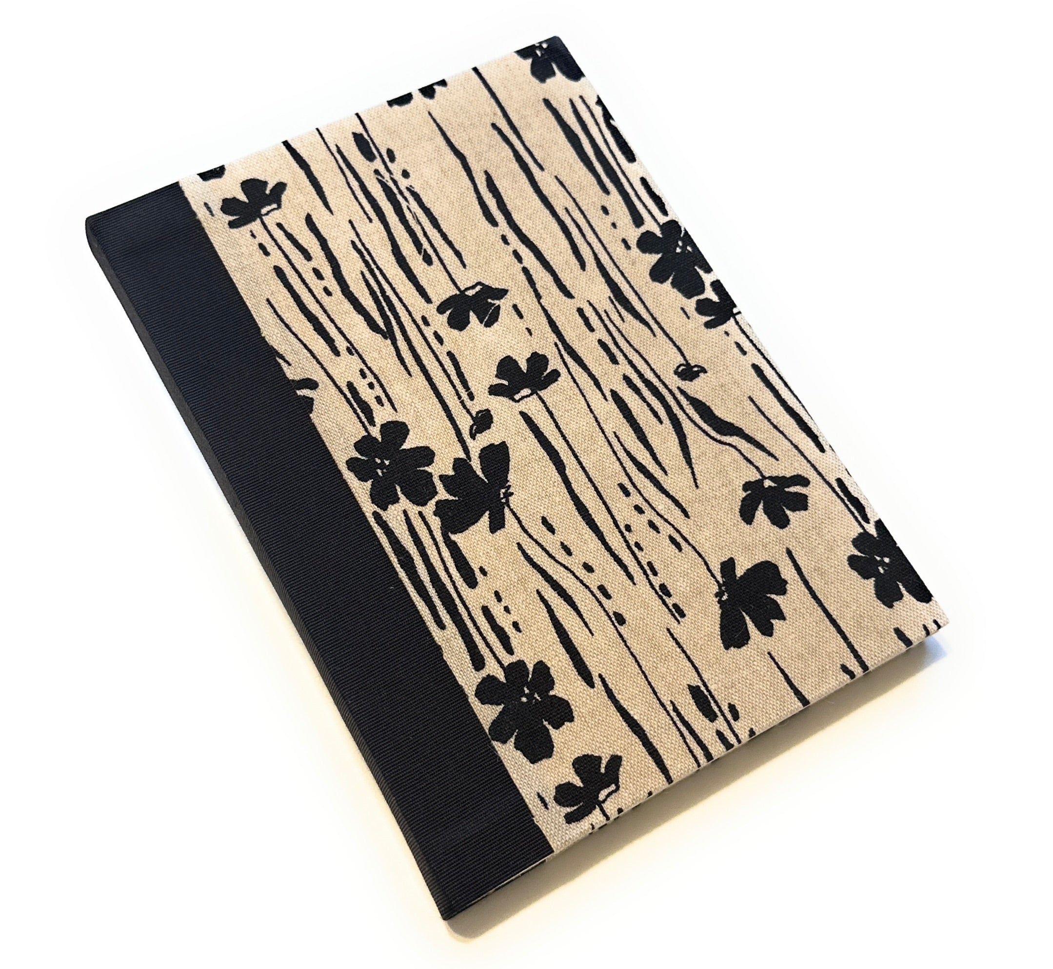 This image shows a handmade book with a beige fabric cover featuring a minimalist black floral pattern. The design includes various black flowers and vertical lines that create a natural, organic look. The spine is covered with solid black fabric, providing a striking contrast to the patterned cover. The book is skillfully crafted, showcasing a clean and professional finish.