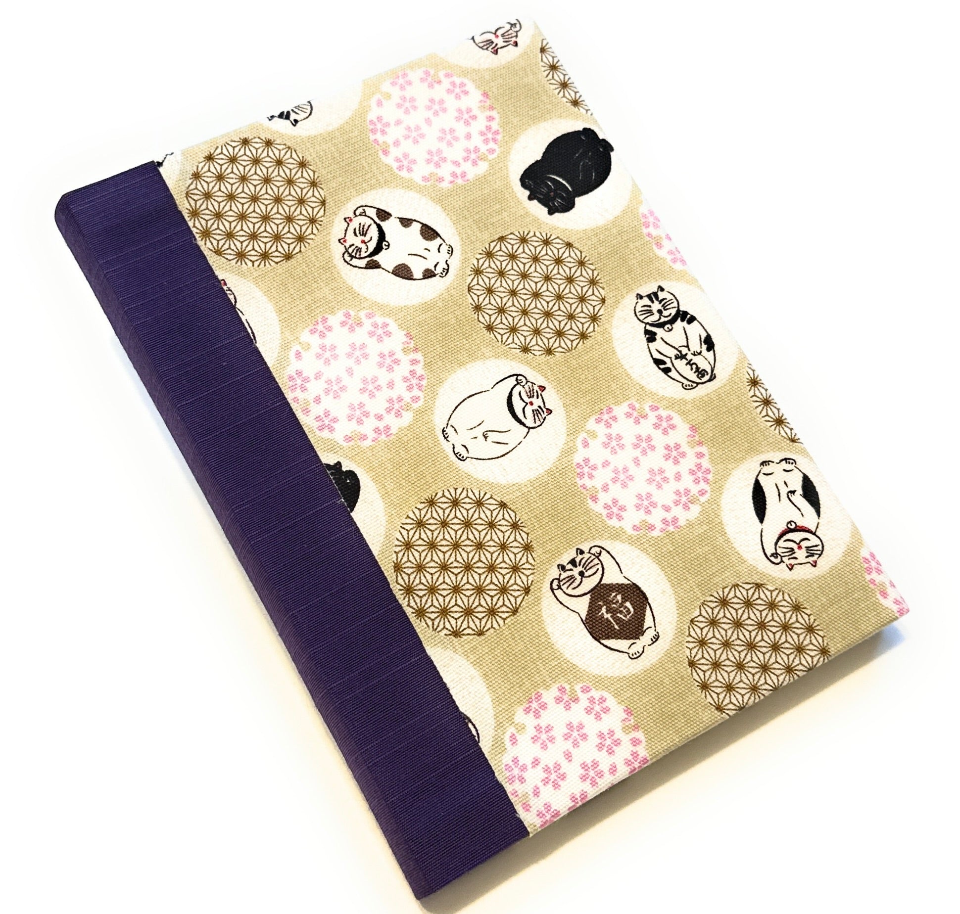 This image shows a handmade book with a beige fabric cover featuring a whimsical design. The cover is adorned with various circular patterns, each containing playful illustrations of cats in different poses. Some circles have floral patterns in pink and brown. The spine is covered with a deep purple fabric, providing a bold contrast to the lighter, playful cover design. The book is expertly crafted, demonstrating a clean and polished finish.