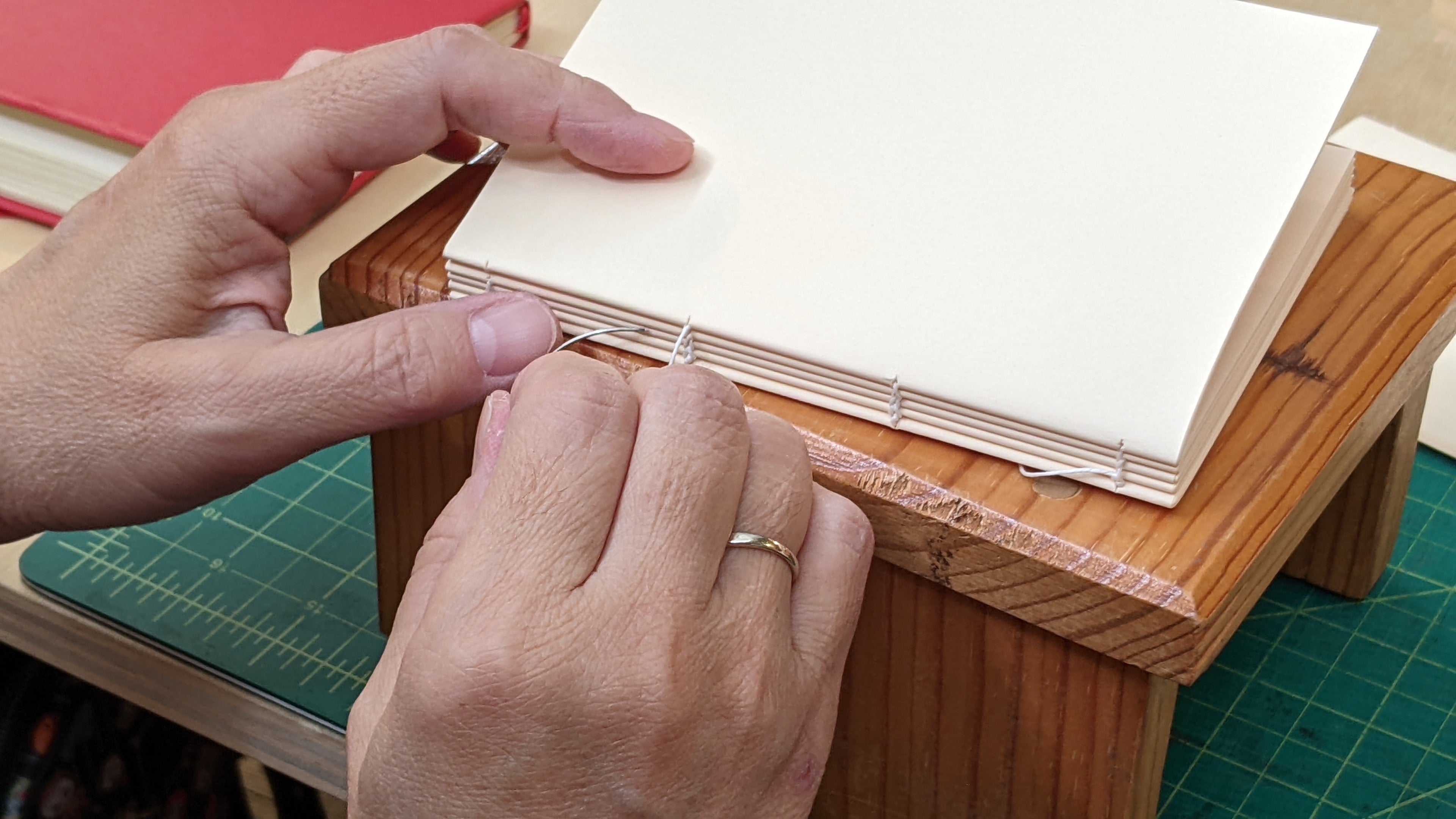 A close-up view of hands engaged in the bookbinding process. The person is stitching the signatures of a book together using a needle and thread, with the signatures neatly aligned on a wooden support. The background includes a green cutting mat and a red book, indicating a workspace dedicated to bookbinding. The image highlights the careful craftsmanship and attention to detail involved in the traditional art of bookbinding.