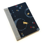 This image shows a handmade book with a dark fabric cover. The fabric has a playful design featuring light cream cats and circular patterns in various shades of blue. There are also small red and yellow flowers scattered across the cover. The spine is covered with a light grey fabric, adding a contrasting element to the overall design. The book appears to be well-crafted, with clean lines and a professional finish.