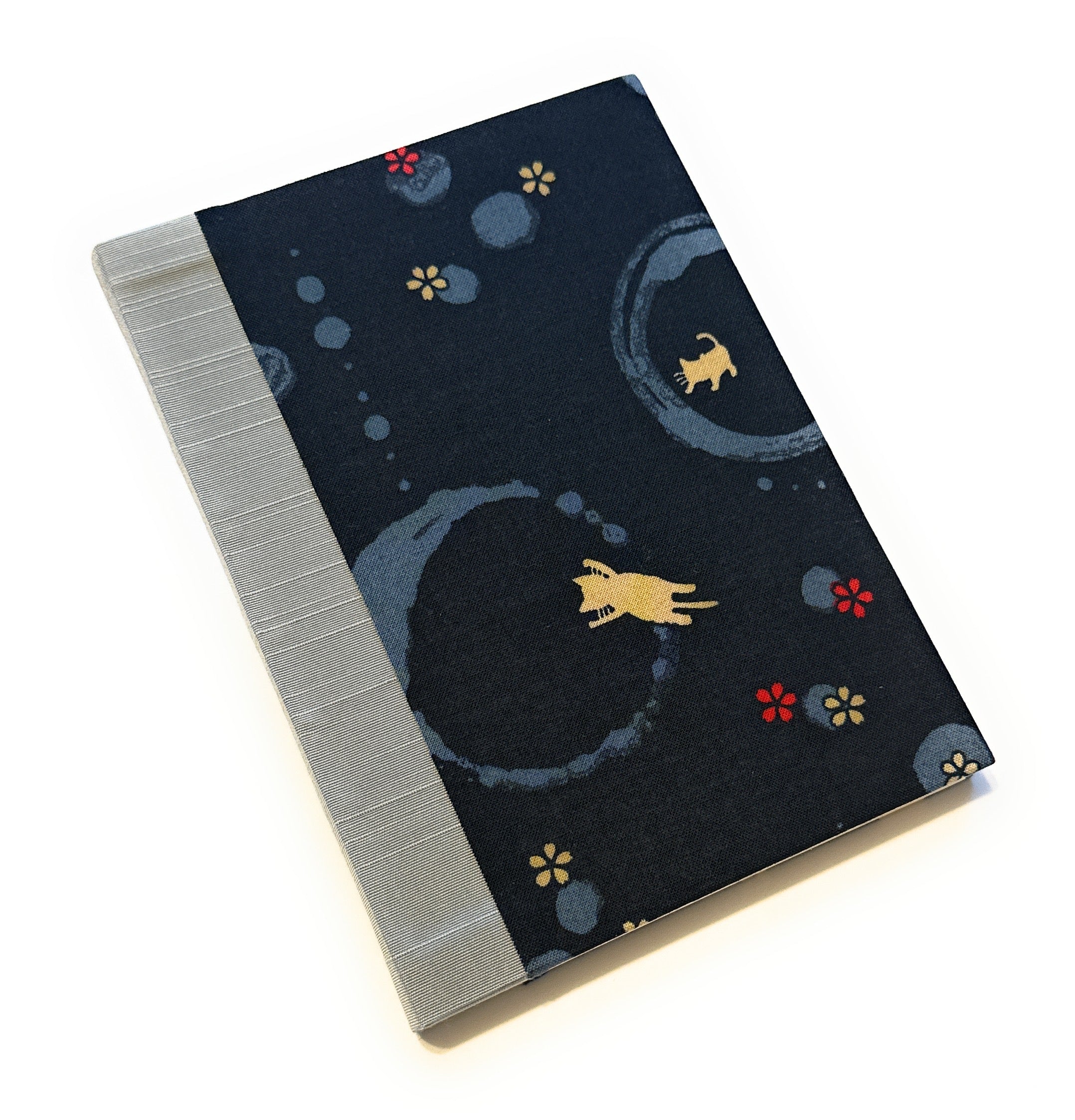 This image shows a handmade book with a dark fabric cover. The fabric has a playful design featuring light cream cats and circular patterns in various shades of blue. There are also small red and yellow flowers scattered across the cover. The spine is covered with a light grey fabric, adding a contrasting element to the overall design. The book appears to be well-crafted, with clean lines and a professional finish.