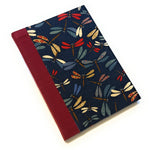 This image shows a handmade book with a dark blue fabric cover adorned with a colorful pattern of dragonflies. The dragonflies are depicted in various shades of red, blue, green, yellow, and white, with intricate designs on their wings. The spine is covered with a rich red fabric that contrasts nicely with the dark blue cover. The book is beautifully crafted, exhibiting a clean and professional finish.