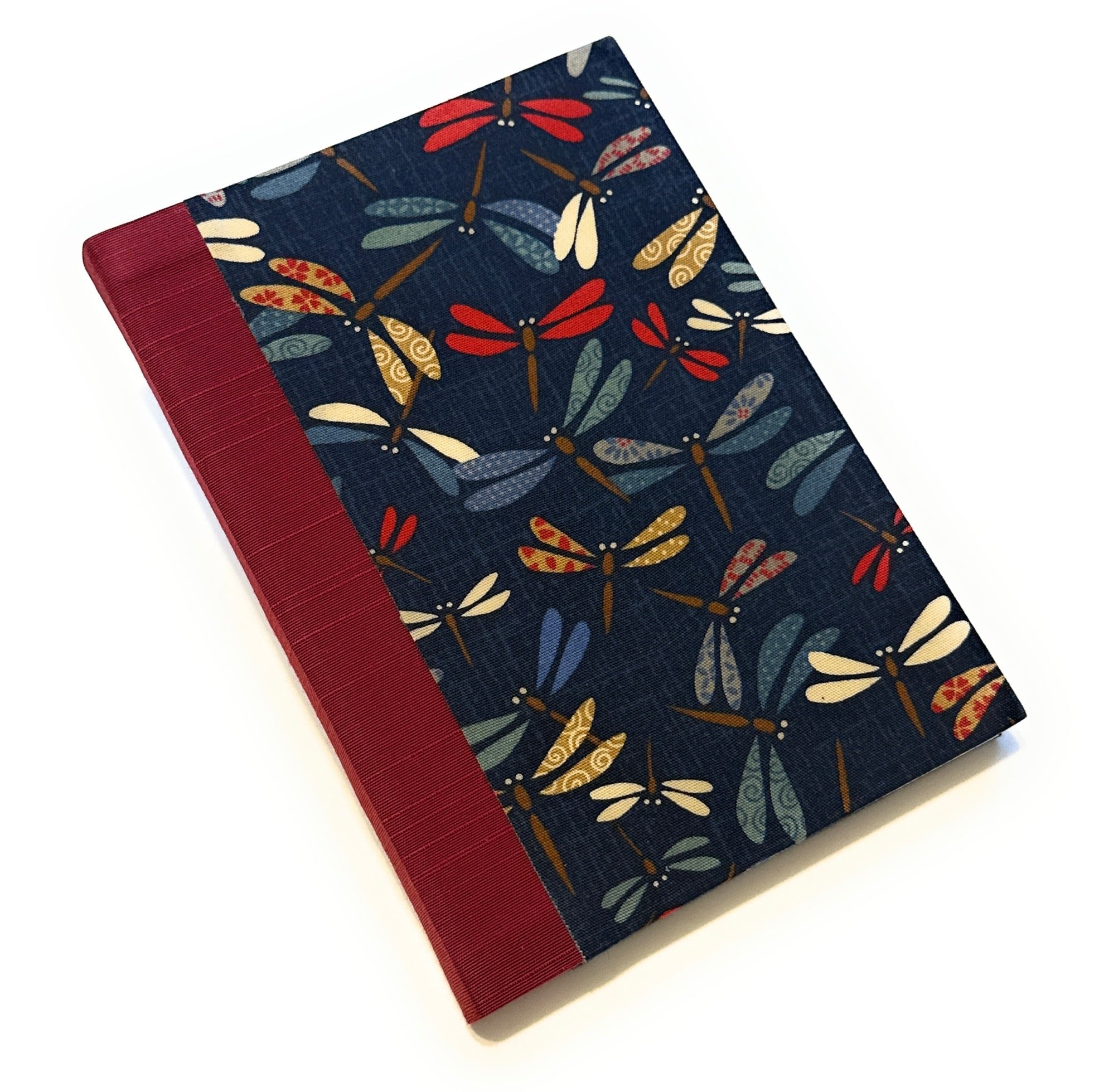 This image shows a handmade book with a dark blue fabric cover adorned with a colorful pattern of dragonflies. The dragonflies are depicted in various shades of red, blue, green, yellow, and white, with intricate designs on their wings. The spine is covered with a rich red fabric that contrasts nicely with the dark blue cover. The book is beautifully crafted, exhibiting a clean and professional finish.