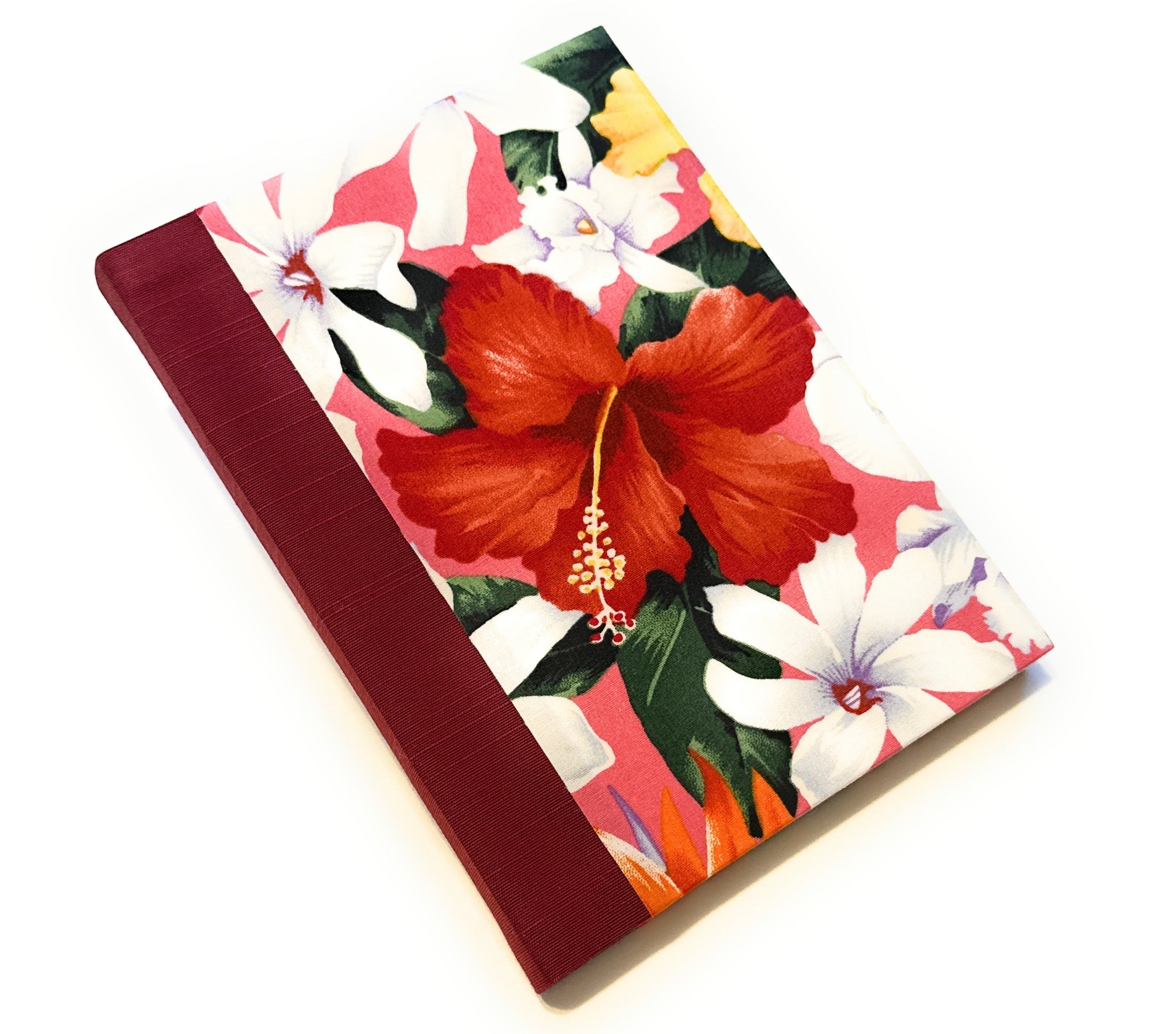 This image shows a handmade book with a vibrant, tropical fabric cover. The cover features large, colorful flowers, including a prominent red hibiscus, Tahitian gardenias, orchids, and other tropical blooms set against a pink background. The spine is covered with a deep red fabric that complements the bright floral design. The book is expertly crafted, exhibiting a clean and polished finish.