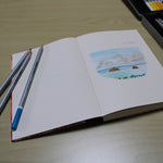 An open sketchbook lies on a wooden table, featuring a colored pencil drawing of two small islands with palm trees in the foreground and a cloudy sky above. Three colored pencils are placed across the left page, and a box of various colored pencils is visible in the background. The sketchbook captures the tranquil scene of the islands, showcasing an artistic moment and the tools used for creating the artwork. The overall setting is calm and conducive to creativity.