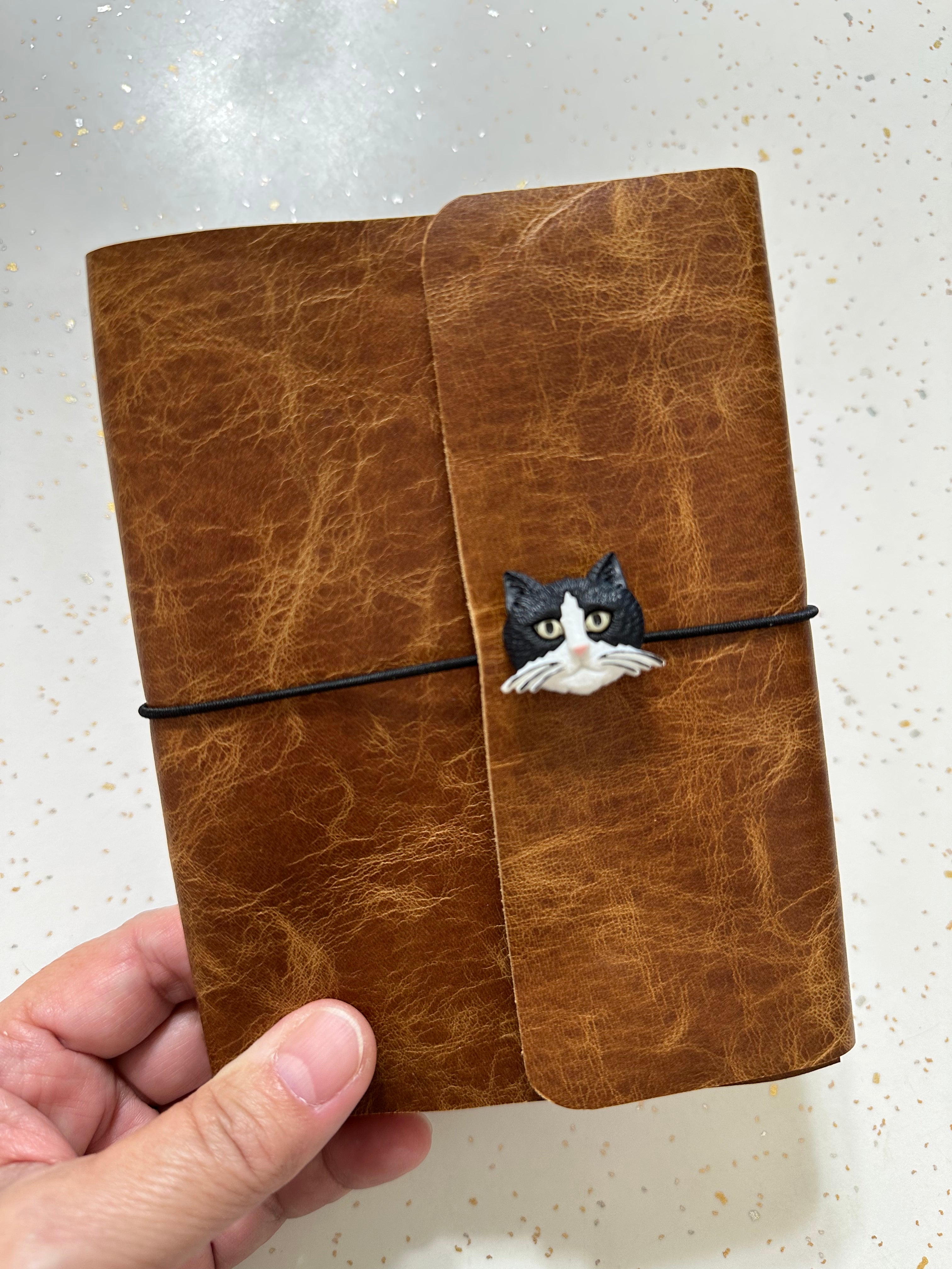 Brown leather notebook with a decorative cat button held by a hand on a light background