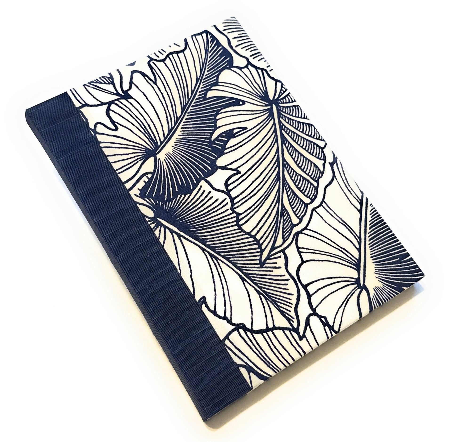 This image shows a handmade book with a white fabric cover featuring a bold, dark blue leaf pattern. The design consists of large, intricately detailed leaves that create a striking visual contrast against the white background. The spine is covered with solid dark blue fabric, complementing the cover's design. The book is skillfully crafted, showcasing a clean and professional finish.