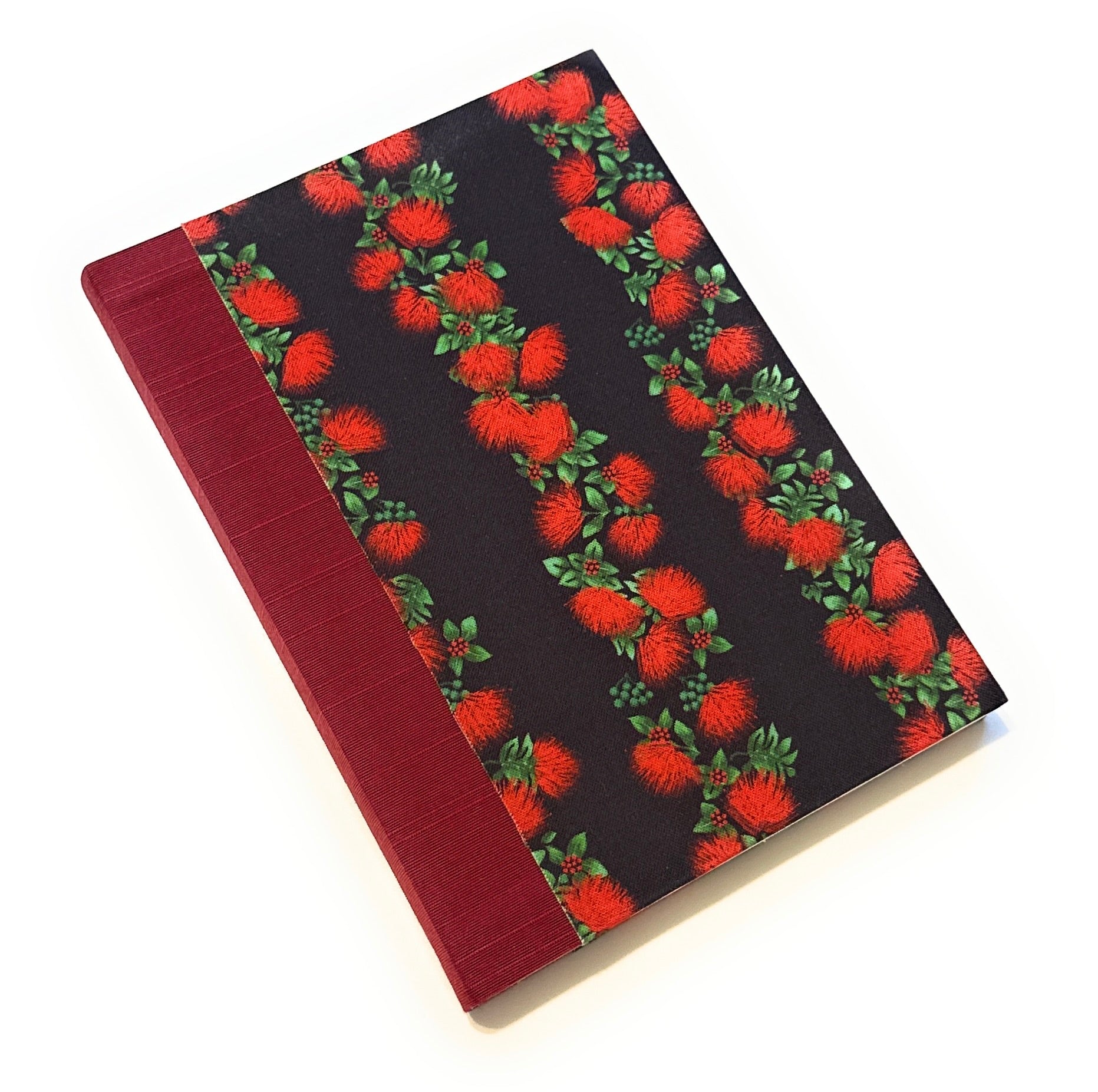 This image shows a handmade book with a black fabric cover featuring a pattern of bright red ʻŌhiʻa flowers and green leaves. The flowers are arranged in vertical stripes, creating a striking contrast against the black background. The spine is covered with a rich, deep red fabric, complementing the red flowers on the cover. The book is meticulously crafted, exhibiting a clean and professional finish.