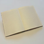 An open journal with cream-colored lined pages lies on a clean, white surface. The journal pages are blank, awaiting written content. The simple and elegant design of the journal highlights its usability for note-taking, journaling, or creative writing. The soft lines on the pages provide a guide for neat handwriting.