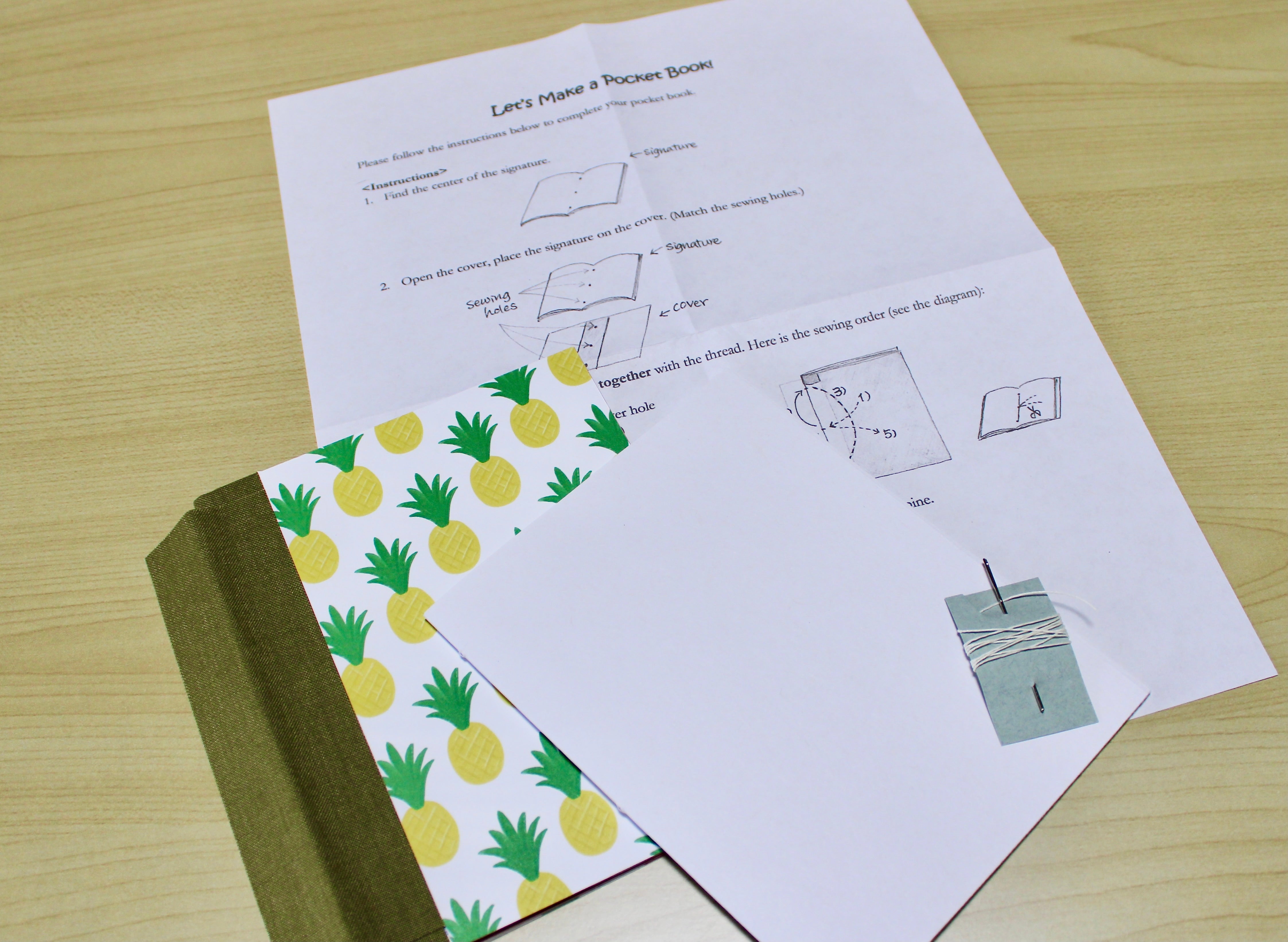 A DIY bookmaking kit featuring a handcrafted pocket journal with a charming yellow pineapple cover and an olive green book cloth spine. The kit includes step-by-step illustrated instructions, sewing thread, a needle, and premium blank paper, making it perfect for beginners and craft enthusiasts who want to create their own personalized booklet.