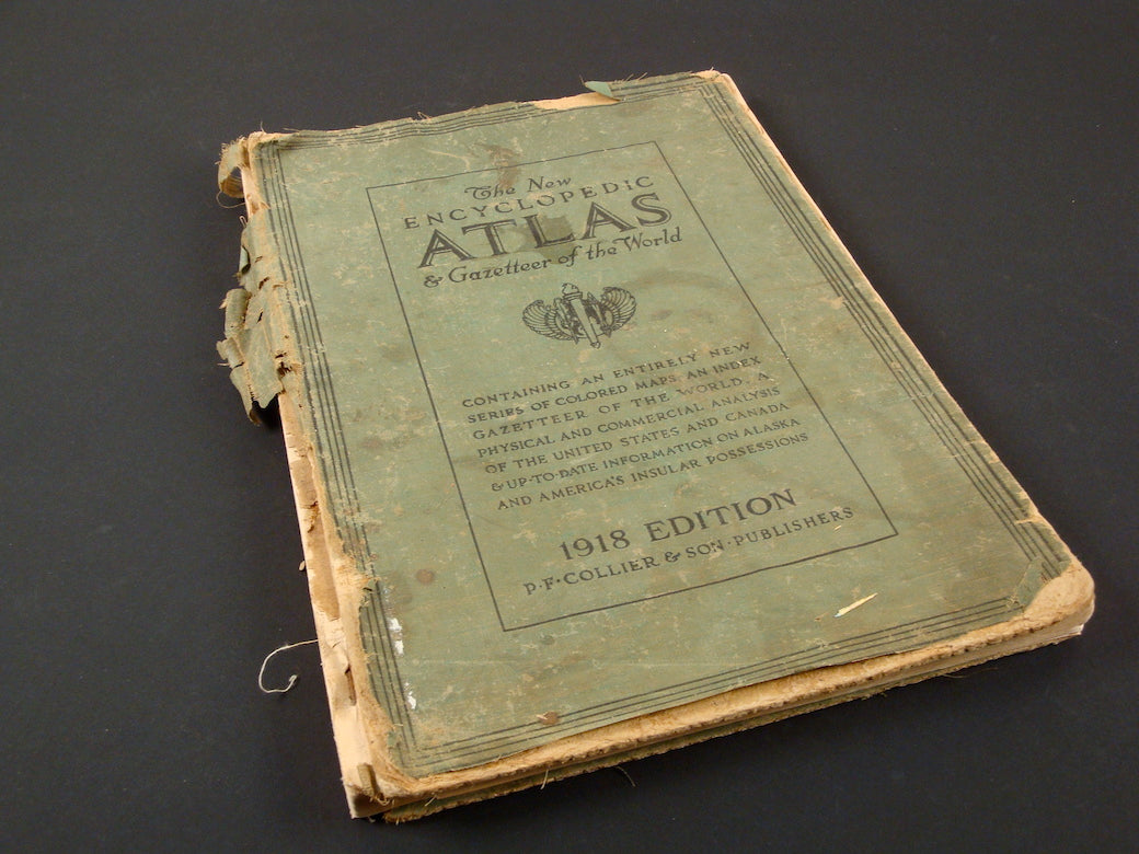 An old, worn book titled “The New Encyclopedic Atlas & Gazetteer of the World” lies on a dark surface. The book, published in 1918 by P.F. Collier & Son, has a green cover that is heavily frayed and tattered along the edges and spine. The text and decorative elements on the cover are faded, indicating significant age and use. Despite its condition, the book retains a sense of historical value and intrigue, hinting at the wealth of information contained within its pages.