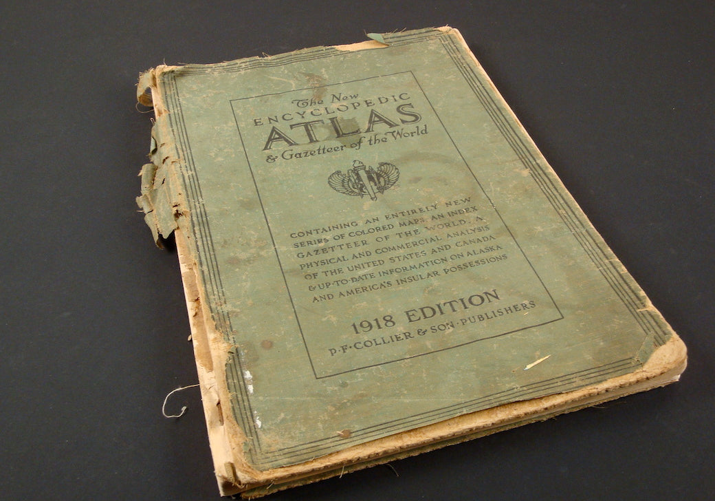 An old, worn book titled “The New Encyclopedic Atlas & Gazetteer of the World” lies on a dark surface. The book, published in 1918 by P.F. Collier & Son, has a green cover that is heavily frayed and tattered along the edges and spine. The text and decorative elements on the cover are faded, indicating significant age and use. Despite its condition, the book retains a sense of historical value and intrigue, hinting at the wealth of information contained within its pages.