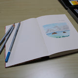 An open handmade journal featuring a colored pencil sketch of a tropical seascape with ocean waves, clouds, and small islands. Three colored pencils rest on the page, while a full set of art supplies sits nearby. This artist’s sketchbook is ideal for drawing, journaling, or travel sketching, offering high-quality paper perfect for creative expression.