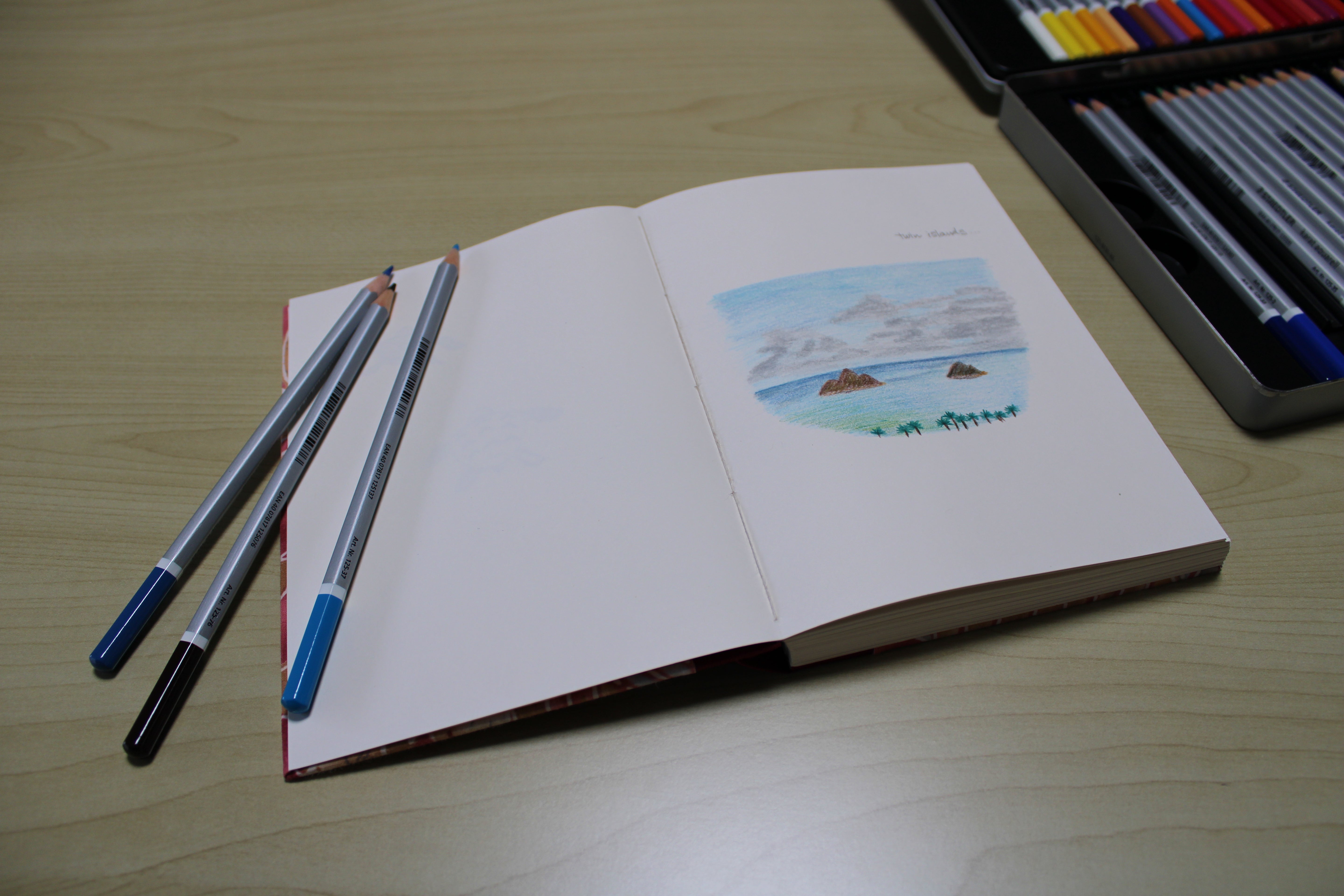An open handmade journal featuring a colored pencil sketch of a tropical seascape with ocean waves, clouds, and small islands. Three colored pencils rest on the page, while a full set of art supplies sits nearby. This artist’s sketchbook is ideal for drawing, journaling, or travel sketching, offering high-quality paper perfect for creative expression.