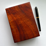 A handcrafted Koa wood Coptic journal with a smooth, polished finish, highlighting the deep reddish-brown grain patterns. A black and gold pen is placed beside it for scale.