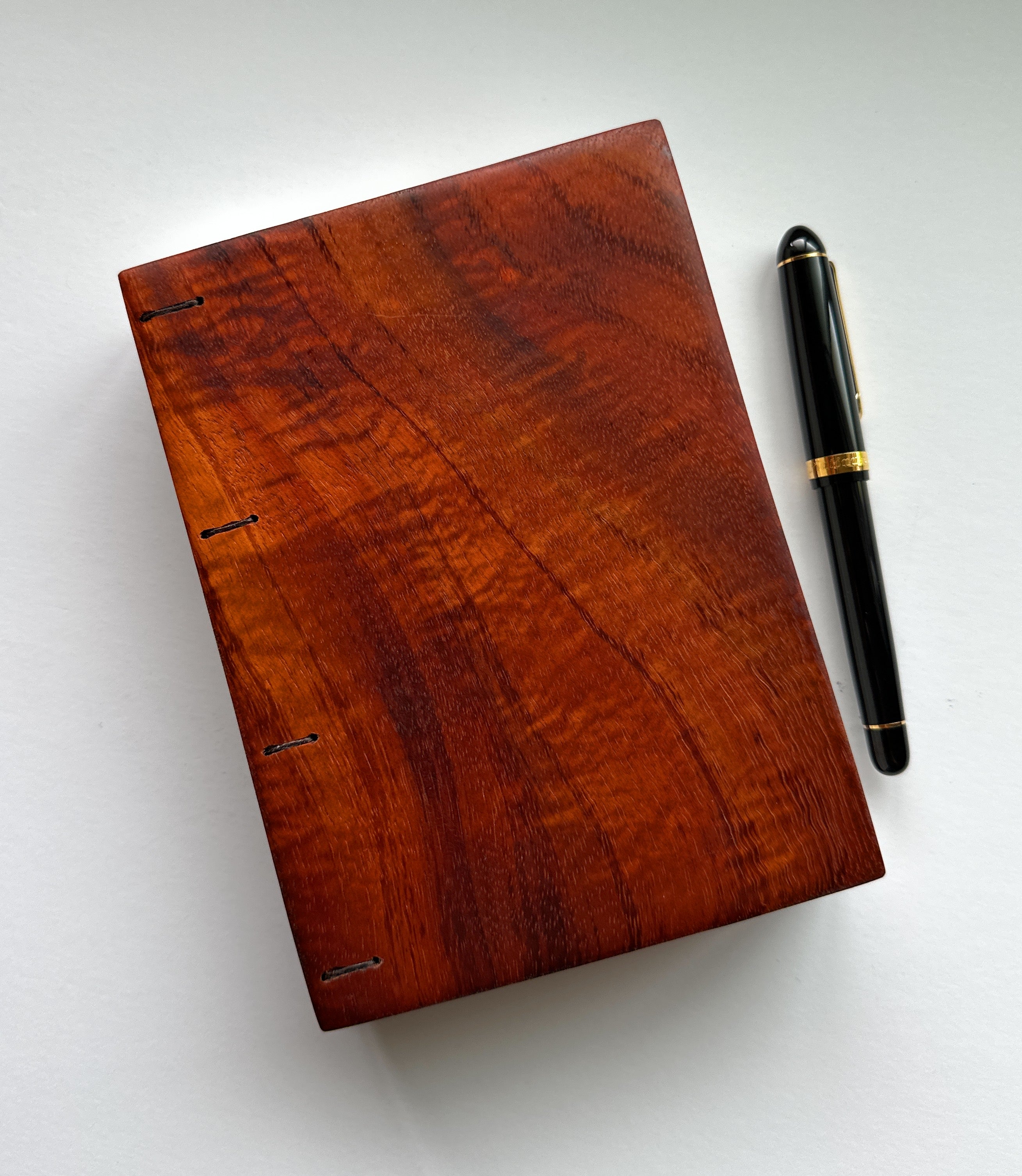 A handcrafted Koa wood Coptic journal with a smooth, polished finish, highlighting the deep reddish-brown grain patterns. A black and gold pen is placed beside it for scale.