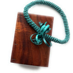 A handcrafted Koa wood Coptic journal featuring a smooth, polished finish. A teal ribbon lei with intricate woven details and a decorative bow is draped over the journal.