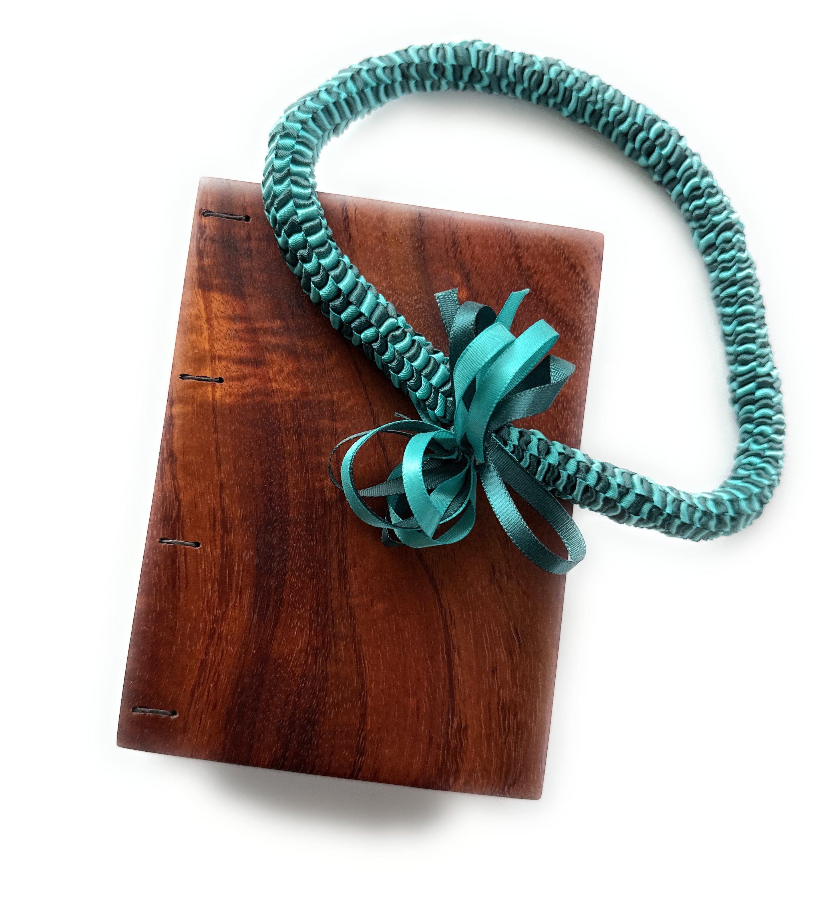 A handcrafted Koa wood Coptic journal featuring a smooth, polished finish. A teal ribbon lei with intricate woven details and a decorative bow is draped over the journal.