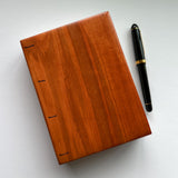 A handmade Lychee wood Coptic journal with a smooth, polished wooden cover in a warm, reddish-brown hue. The minimalist design highlights the natural wood grain. A luxurious black and gold fountain pen rests beside the journal, emphasizing its use for writing, sketching, or journaling. The flat-lay binding makes it an ideal choice for creatives seeking a high-quality, artisanal notebook.