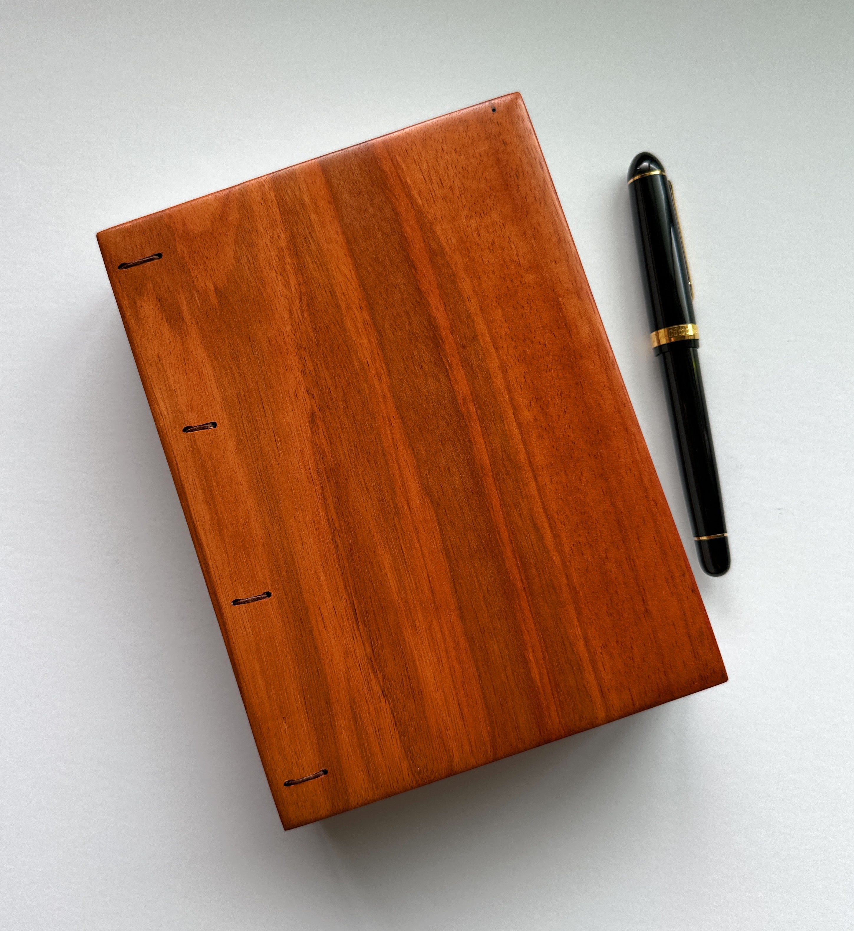 A handmade Lychee wood Coptic journal with a smooth, polished wooden cover in a warm, reddish-brown hue. The minimalist design highlights the natural wood grain. A luxurious black and gold fountain pen rests beside the journal, emphasizing its use for writing, sketching, or journaling. The flat-lay binding makes it an ideal choice for creatives seeking a high-quality, artisanal notebook.