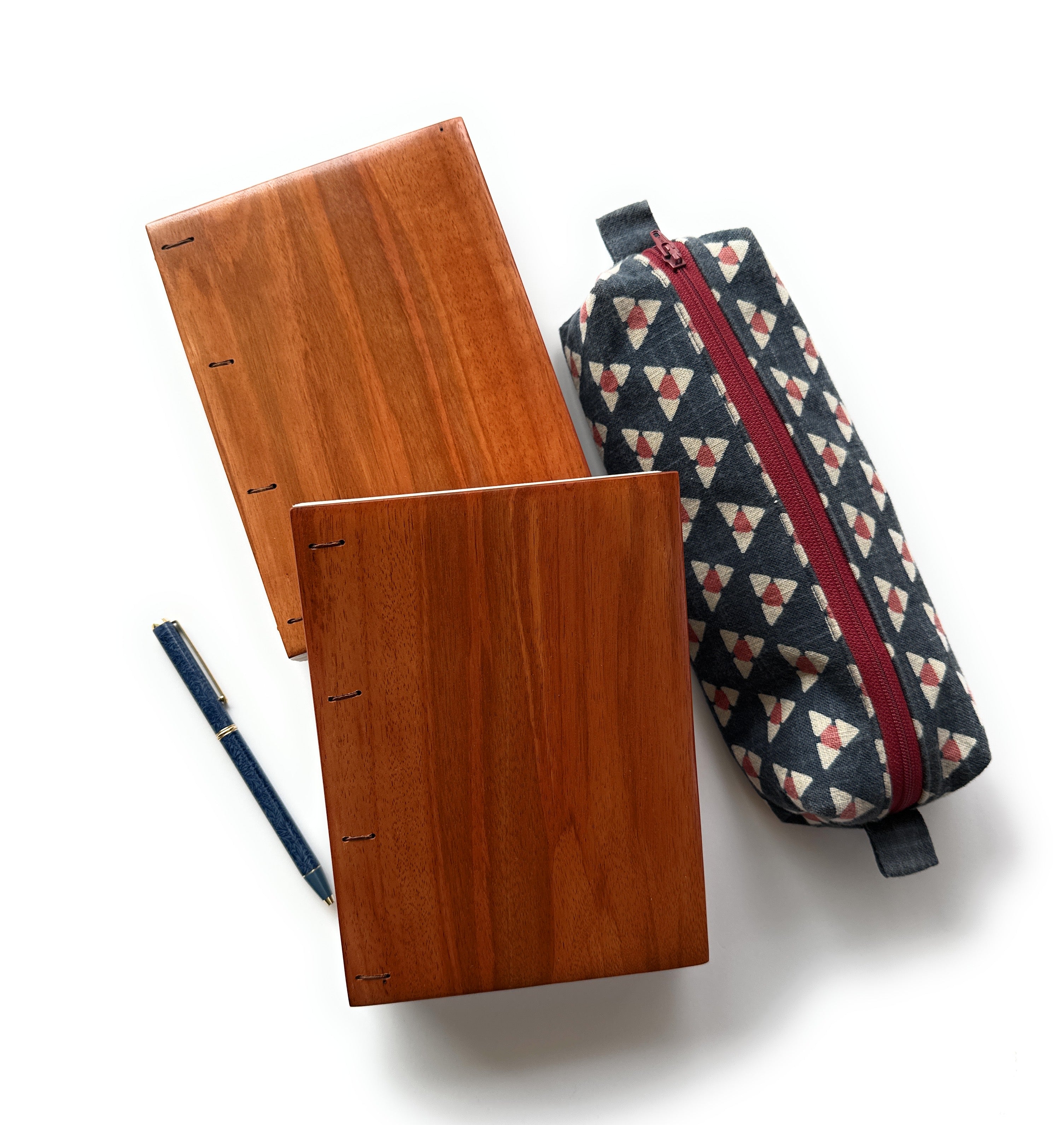 Two handmade Lychee wood Coptic journals with a rich, warm reddish-brown finish and visible Coptic stitch binding, laid flat on a white surface. Accompanied by a blue fabric-covered fountain pen and a geometric-patterned pencil case with a red zipper, this image showcases a stylish and functional stationery set. The smooth wooden covers highlight the natural grain, making these journals perfect for writing, sketching, or journaling with a timeless, artisanal touch.
